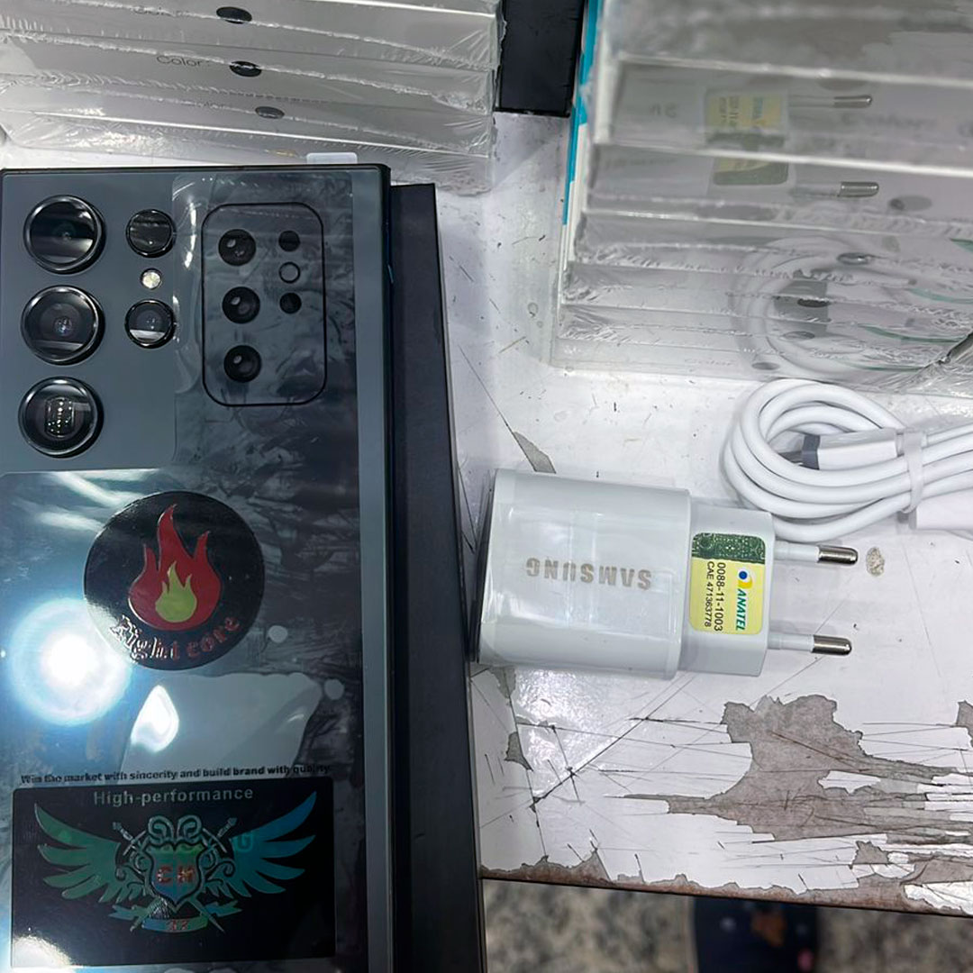 S24 Ultra Goophone Preto com acessórios S24 Ultra Goophone preto sobre mesa com cabo, conector e película.